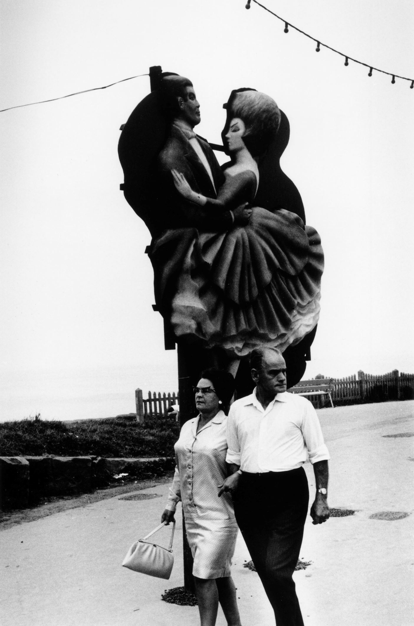 Male and female couple walking in front of a Blackpool Illumination of dancing couple. Included in the exhibition Mass Photography: Blackpool Through The Camera