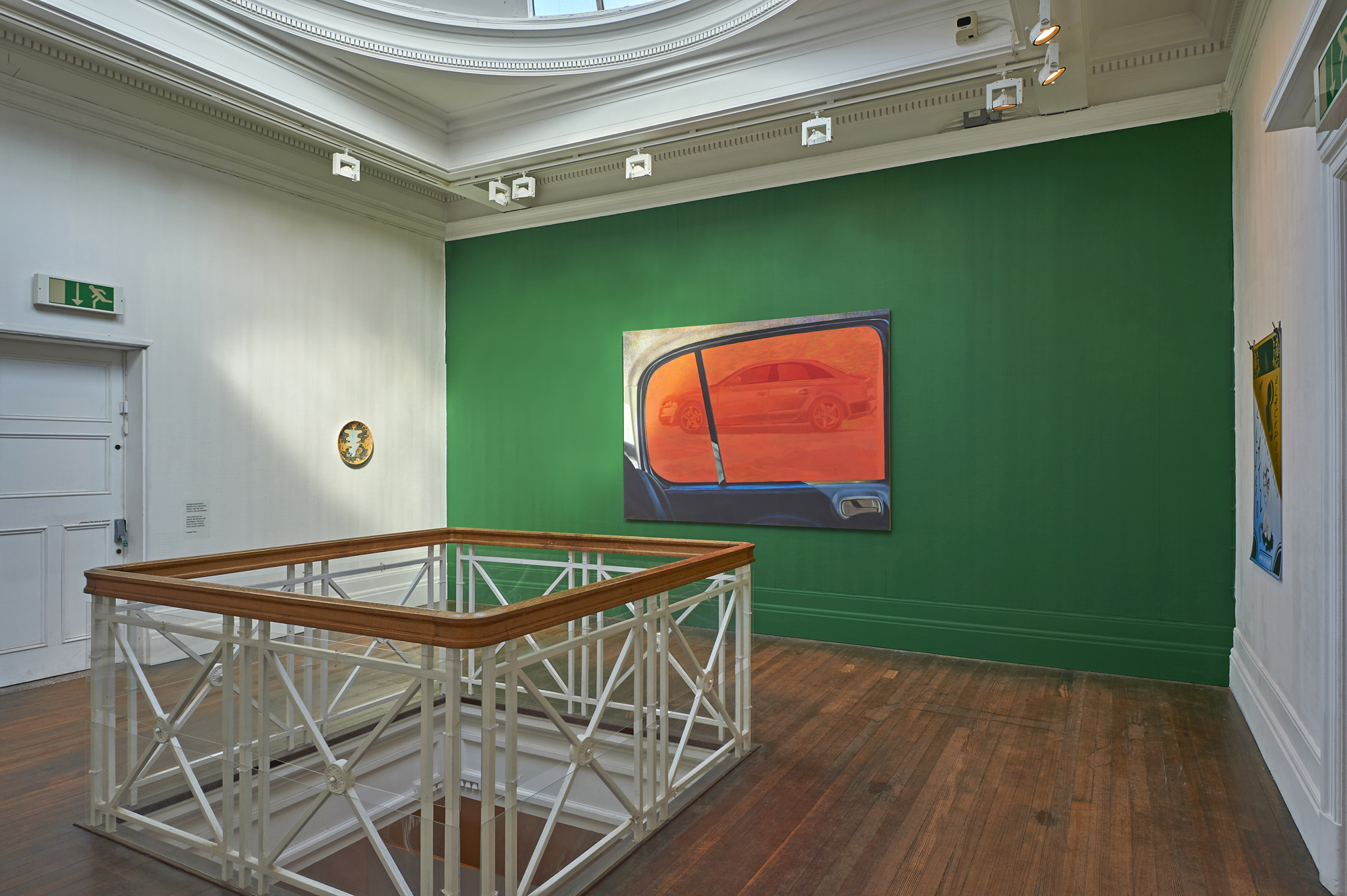 The Grundy Art Gallery’s upstairs rotunda space featuring painting of view from a cars side window, the view is of another car. The painting by artist Alison Katz is displayed against a green background. Included in the exhibition Fig-Futures: Alison Katz.