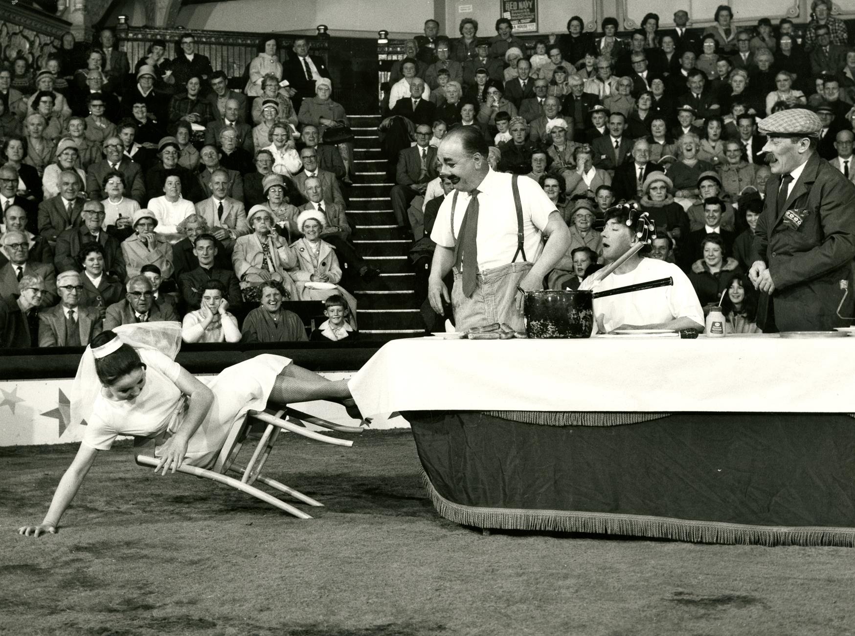 3 clowns including Charlie Cairoli watching a woman in white dress falling off a chair. Photograph included in the exhibition Come One, Come All!: A Celebration Of Blackpool Tower Circus.